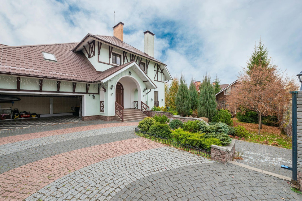 Elegant interlocking driveway with a curved paver design leading to a Tudor-style home surrounded by manicured landscaping and evergreen shrubs.