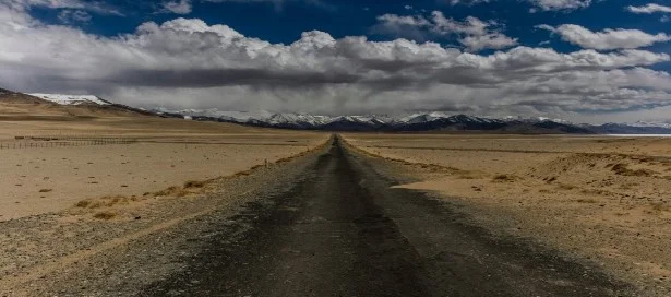 A long, open asphalt road stretching through rugged desert terrain under a bright blue cloudy sky along the Pamir Highway in Tajikistan.